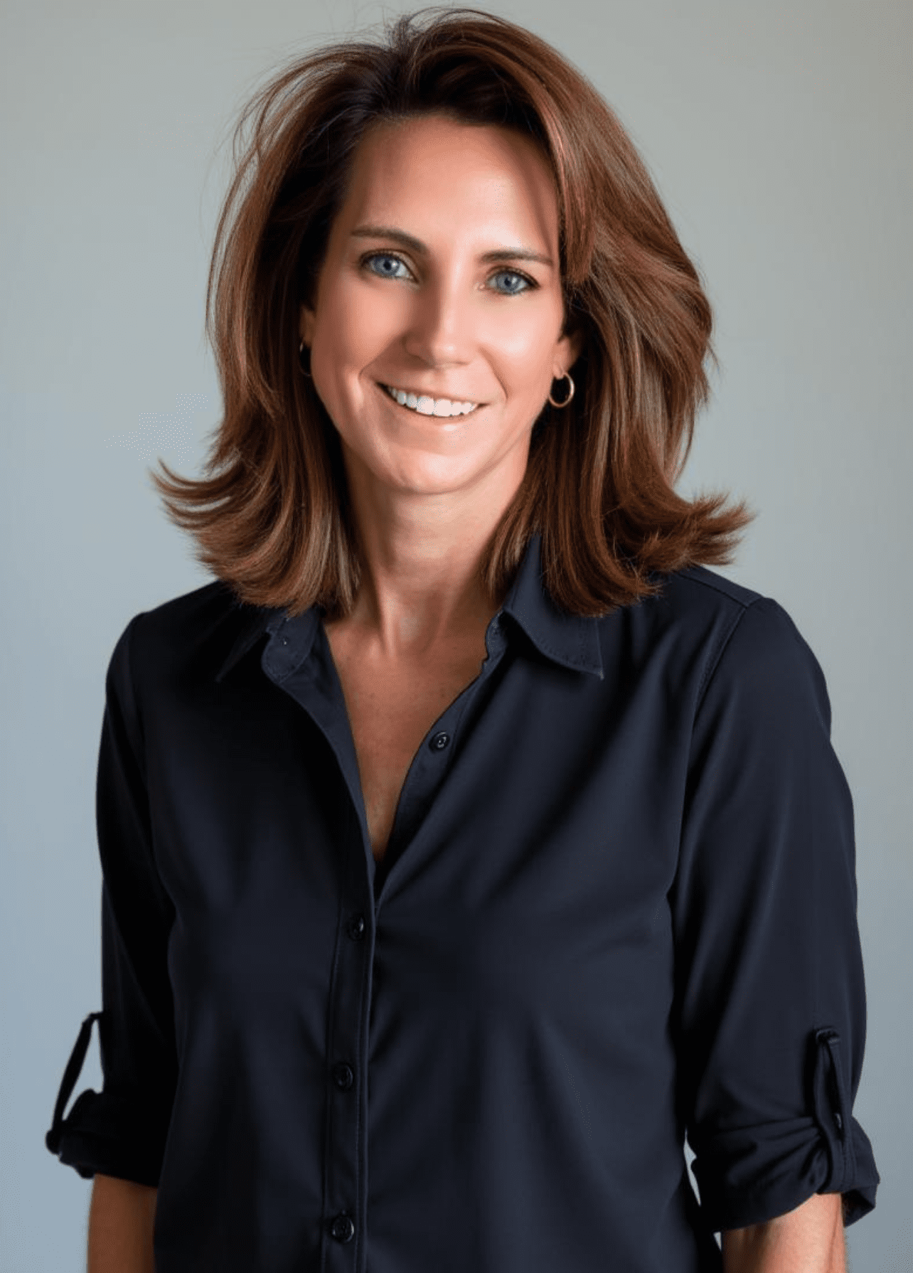 Portrait of a person with shoulder-length light brown hair, wearing a blue shirt and black blazer, looking directly at the camera with a neutral expression.
